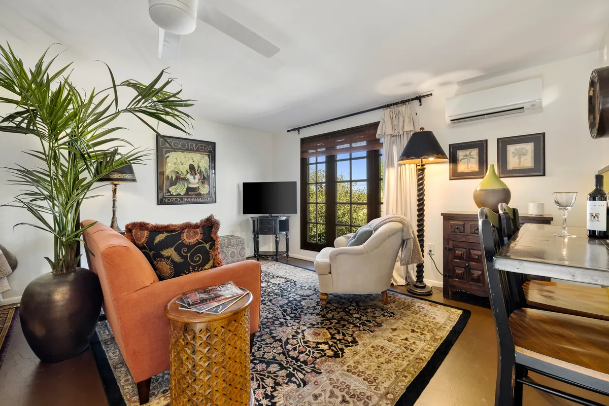 A sunlit living room featuring a vibrant orange sofa, a patterned rug, a dining table, lush potted plant, and a large window that frames the scenic rock formations beyond. The room exudes warmth, comfort, and is designed for both relaxation and gathering with friends or family.