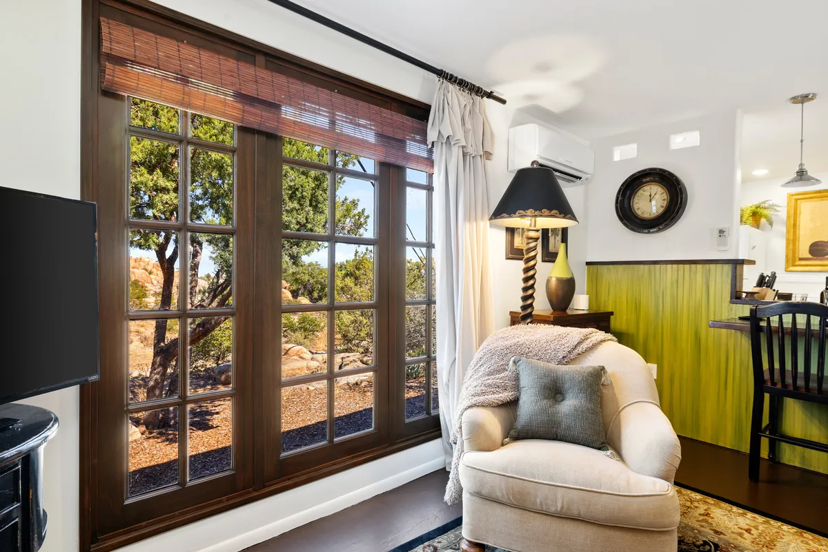 A tranquil reading nook with a large window framed in wood, a plush armchair, decorative lamp, and green accent wall. The window offers peaceful views and bathes the space in natural light, ideal for serene mornings or cozy gatherings.