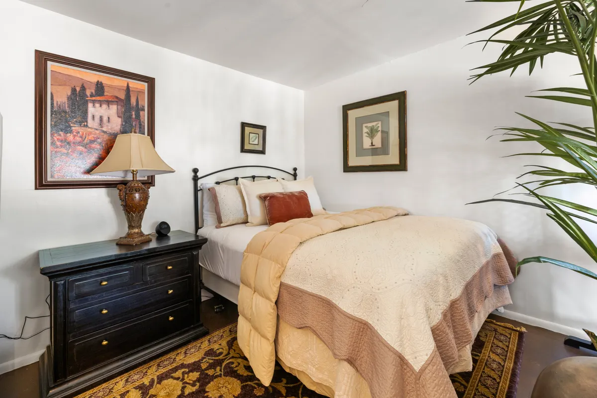 A tranquil bedroom scene featuring a bed with plush decorative bedding, a lamp softly illuminating a nightstand, and calming artwork on the wall. Natural light gently filters into the room, creating a serene and private atmosphere ideal for rest.