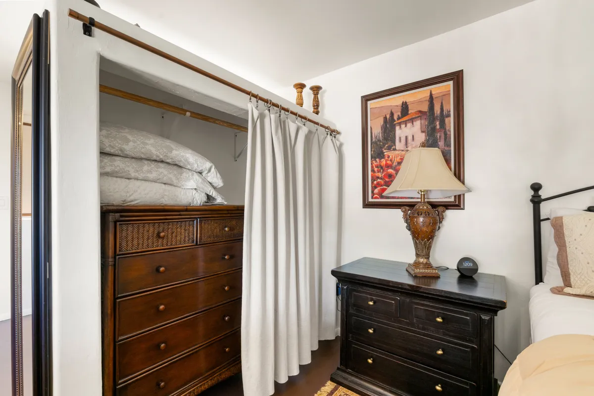 An inviting bedroom corner featuring a dresser, curtained closet, lamp, and a painting. The soft ambient lighting and tasteful furnishings emphasize comfort and a homey, restful vibe.