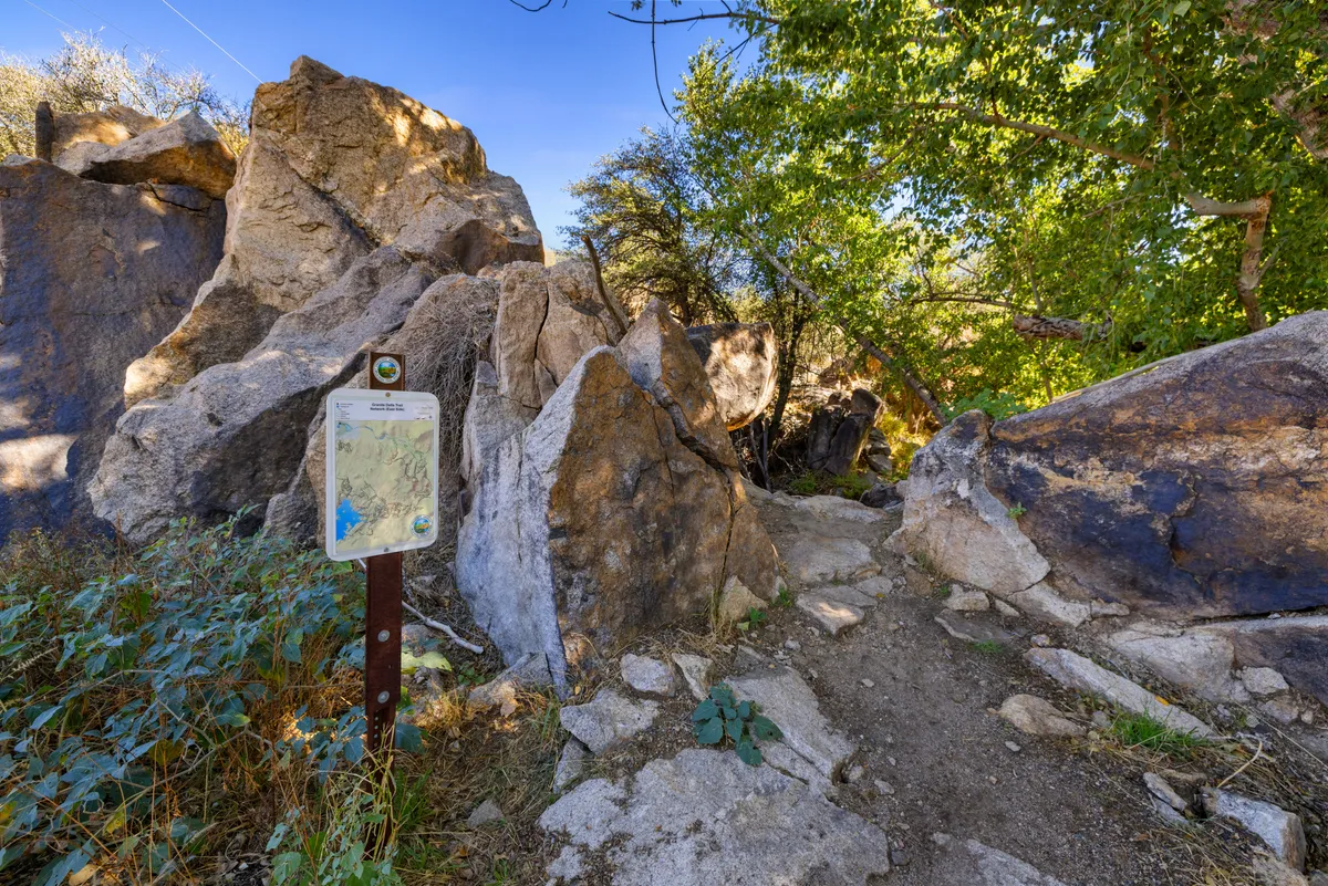 This image showcases a scenic exterior scene featuring large rock formations, a trail sign, a well-trodden pathway, and vibrant surrounding greenery. The atmosphere is both adventurous and tranquil, emphasizing the spirit of exploration and connection to nature.