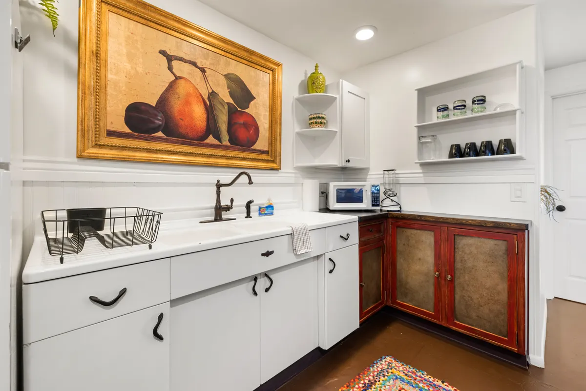 A cozy kitchen scene with a striking fruit painting, antique-style faucet, colorful rug, and wooden cabinets. The setup invites guests to gather, share a meal, or sip coffee together in an intimate, welcoming environment.