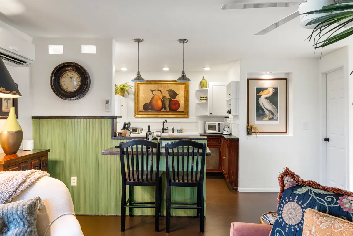 This image showcases a welcoming kitchen with a central island, artwork including a fruit painting, and a decorative chair. The scene captures the essence of a homey kitchen where cooking, entertaining, and socializing take place, with family members enjoying time around the island, possibly preparing meals or sipping wine together.