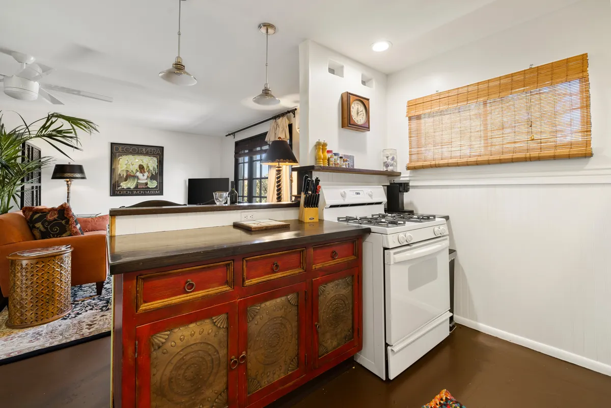 A cozy interior scene with a striking red cabinet adorned with intricate designs, a white gas stove, bamboo window shade, potted plant, and a plush sofa. Large windows allow ample natural light, subtly connecting the interior to the outdoors.