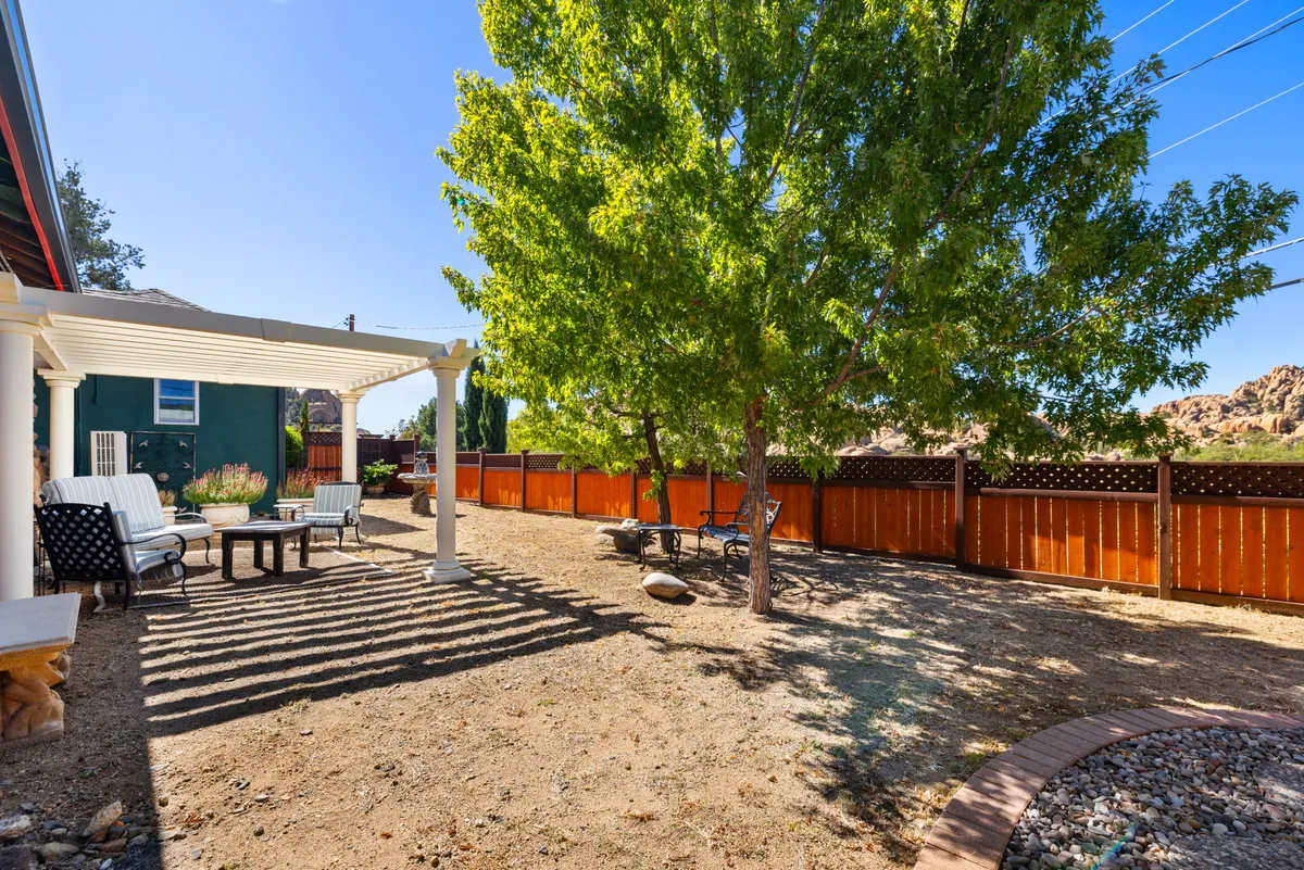 This exterior image captures a tranquil backyard seating area beneath a pergola, with lush greenery and a welcoming fenced yard. The space is designed for outdoor dining and relaxation, exuding an inviting atmosphere that hints at the leisurely pace and outdoor charm of Prescott.