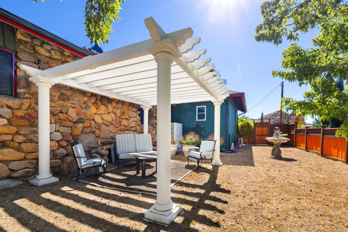 An inviting exterior space featuring a pergola over a stone patio, comfortable seating, a fountain, and a low stone wall. The area is surrounded by lush greenery, offering a perfect spot to enjoy morning coffee or relax in the evening.